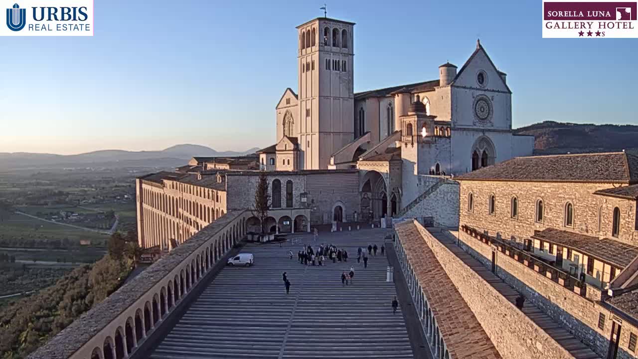 A large stone basilica and adjacent buildings are seen from above on a clear, sunny day, with a valley visible in the background.