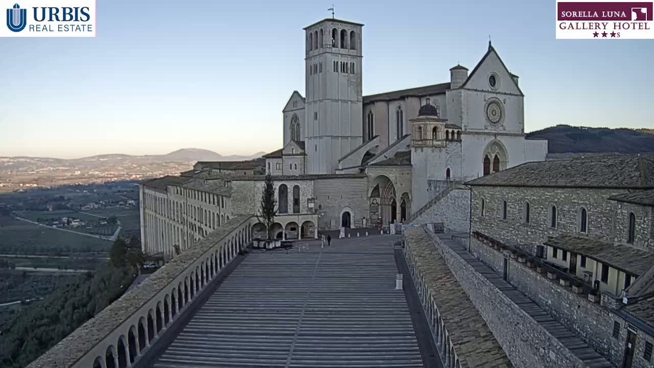 A large stone basilica and adjoining buildings are seen on a clear, sunny day, overlooking a valley.