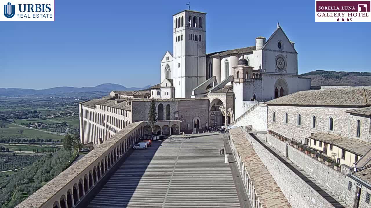 A large, light-colored basilica with a tall bell tower sits atop a hill overlooking a valley on a clear, sunny day.