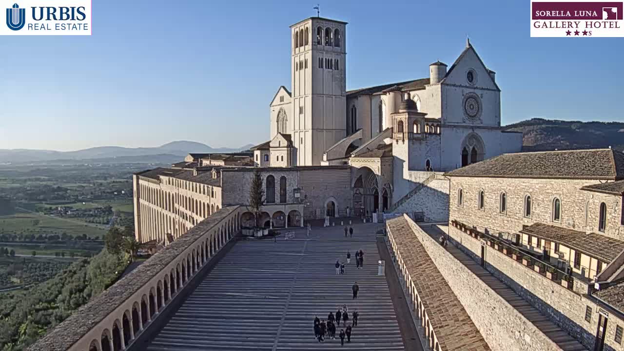 A large stone basilica and adjoining buildings are seen on a clear, sunny day, with a wide plaza in front and a distant view of rolling hills and valleys.