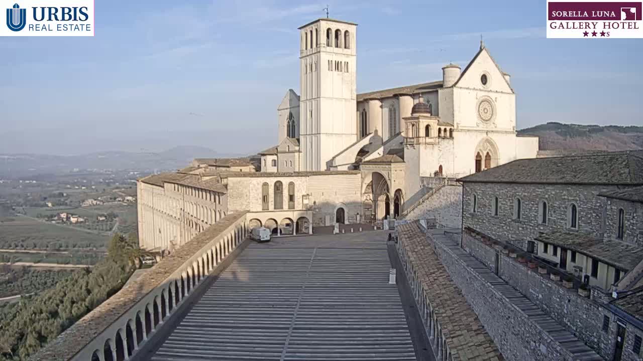 A large, light-colored basilica complex sits atop a hill overlooking a valley on a clear, sunny day.