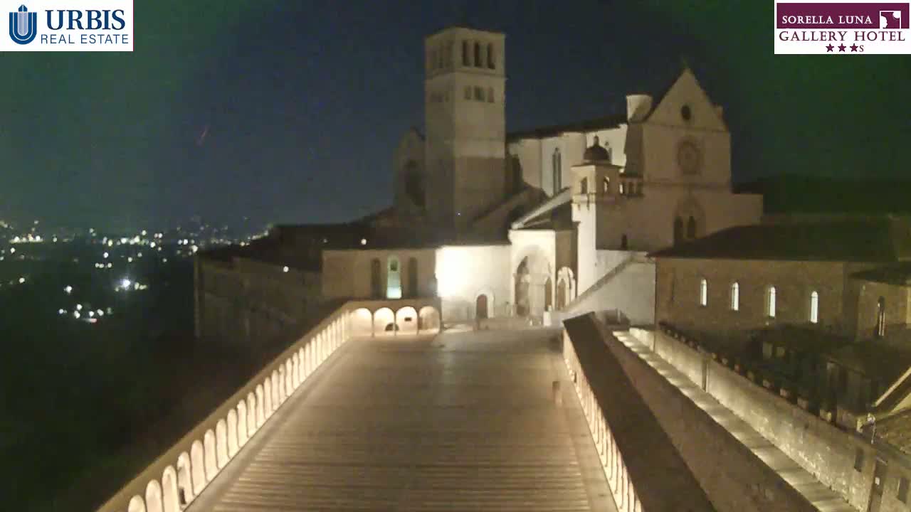 At night, a large, illuminated stone building complex is viewed from above, overlooking a city illuminated by distant lights under a clear, dark sky.