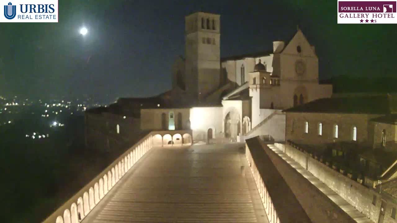 A brightly lit plaza at night, overlooked by a large stone building, under a mostly clear sky with a visible moon.