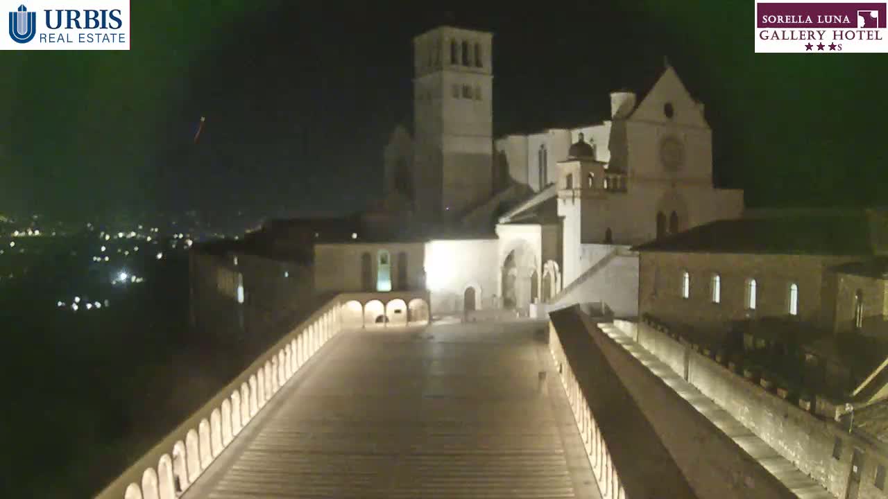 A nighttime view of a large, illuminated stone building complex, possibly a church or monastery, overlooks a city at night.