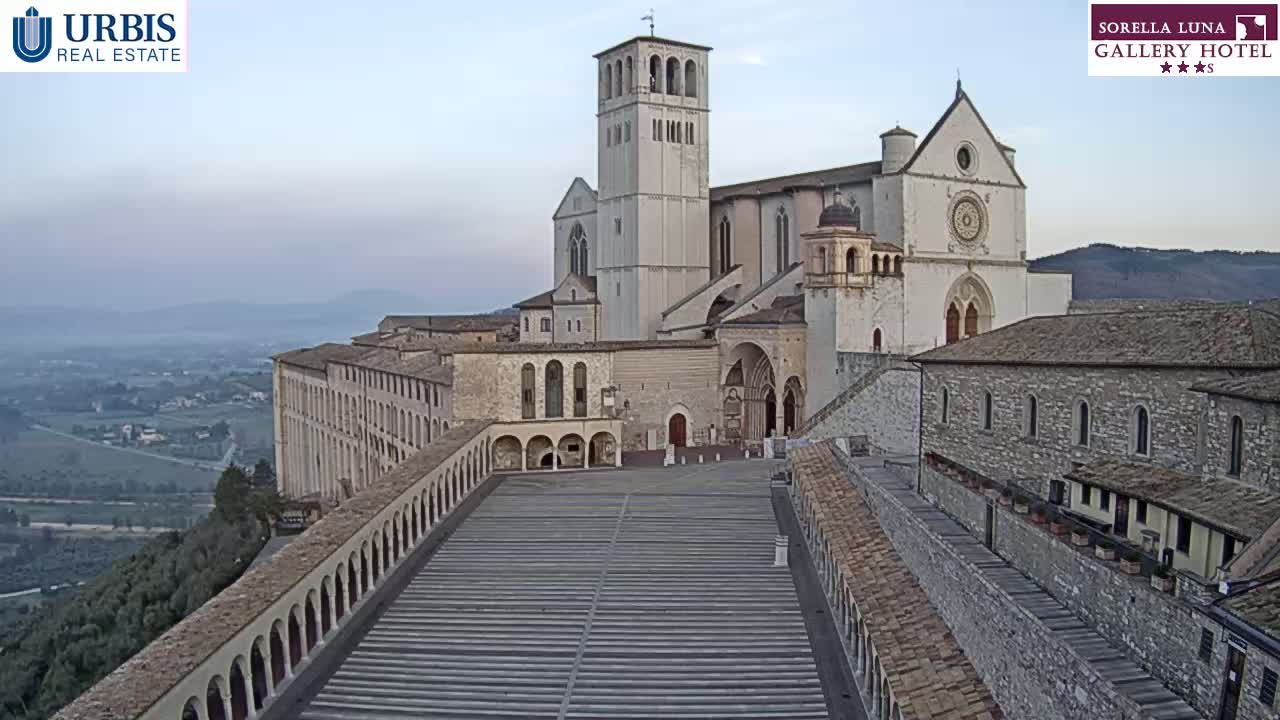 A large stone basilica and adjacent buildings overlook a paved plaza and a valley under a hazy sky.