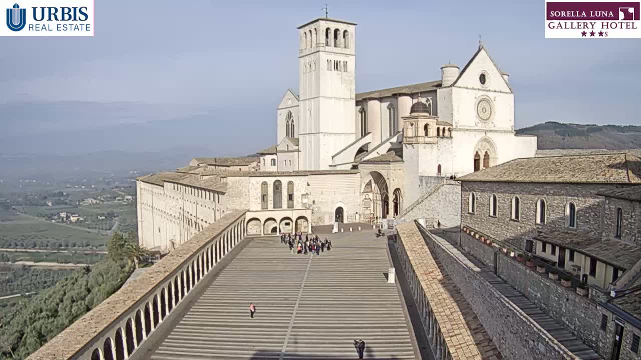 A large, light-colored basilica and adjoining buildings are viewed from above on a sunny day, with a paved plaza in the foreground containing a small group of people.
