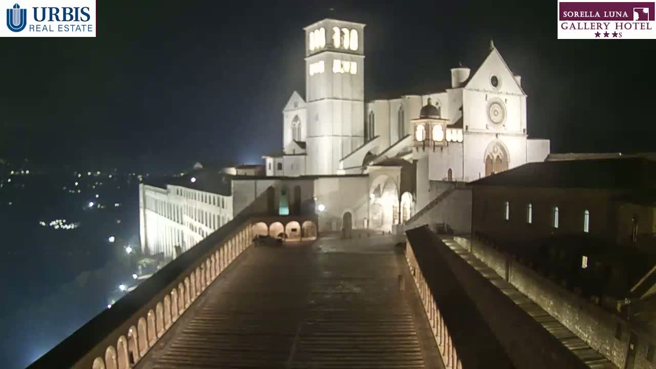 A large, illuminated basilica stands at night, overlooking a long, paved walkway bordered by low walls.