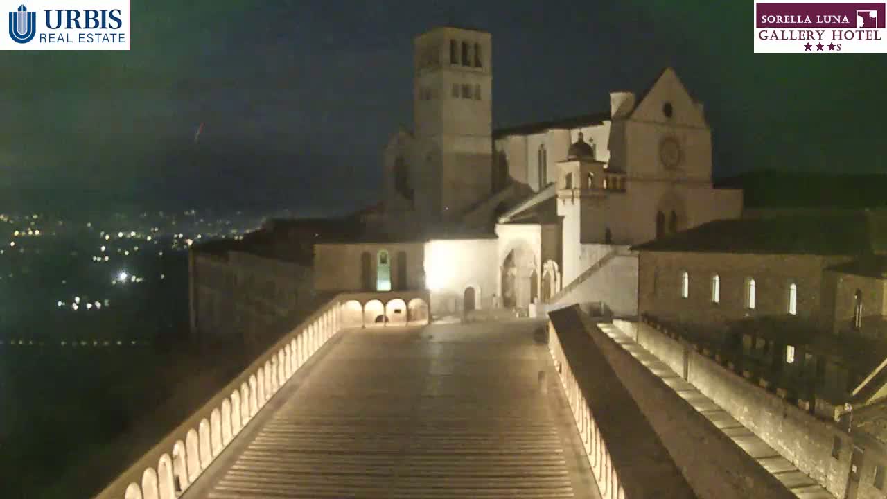A large, illuminated stone building complex sits on a hill at night, viewed from a long, descending walkway, with a city’s lights visible in the distance.