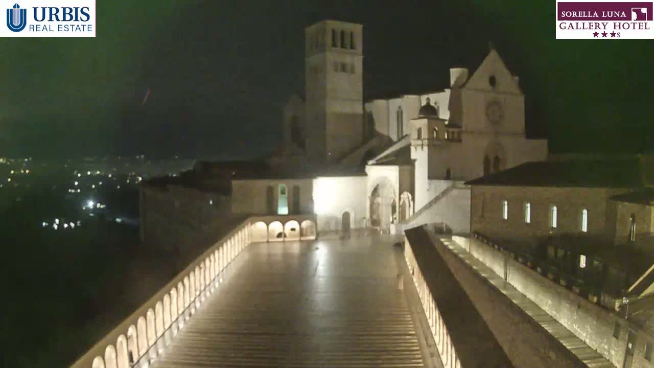 A large, illuminated stone complex, possibly a church or monastery, is seen at night under a dark, possibly overcast sky.