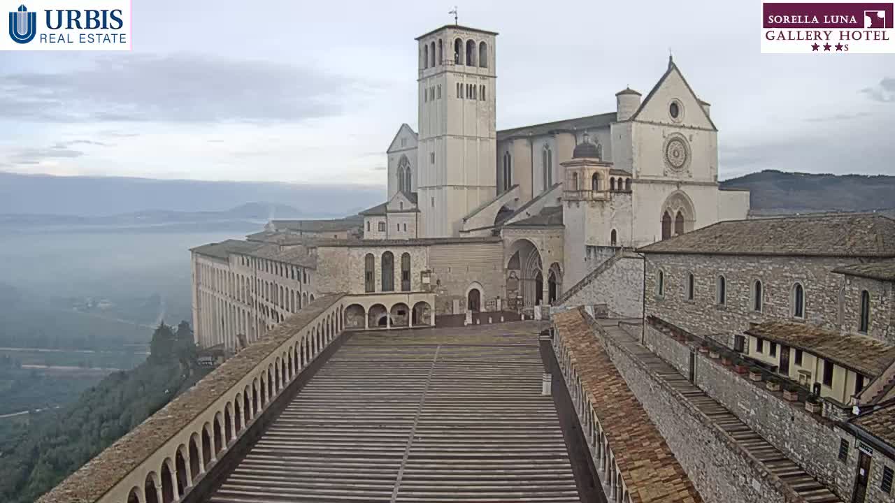 A large, light-colored basilica complex sits atop a hill overlooking a foggy valley under an overcast sky.