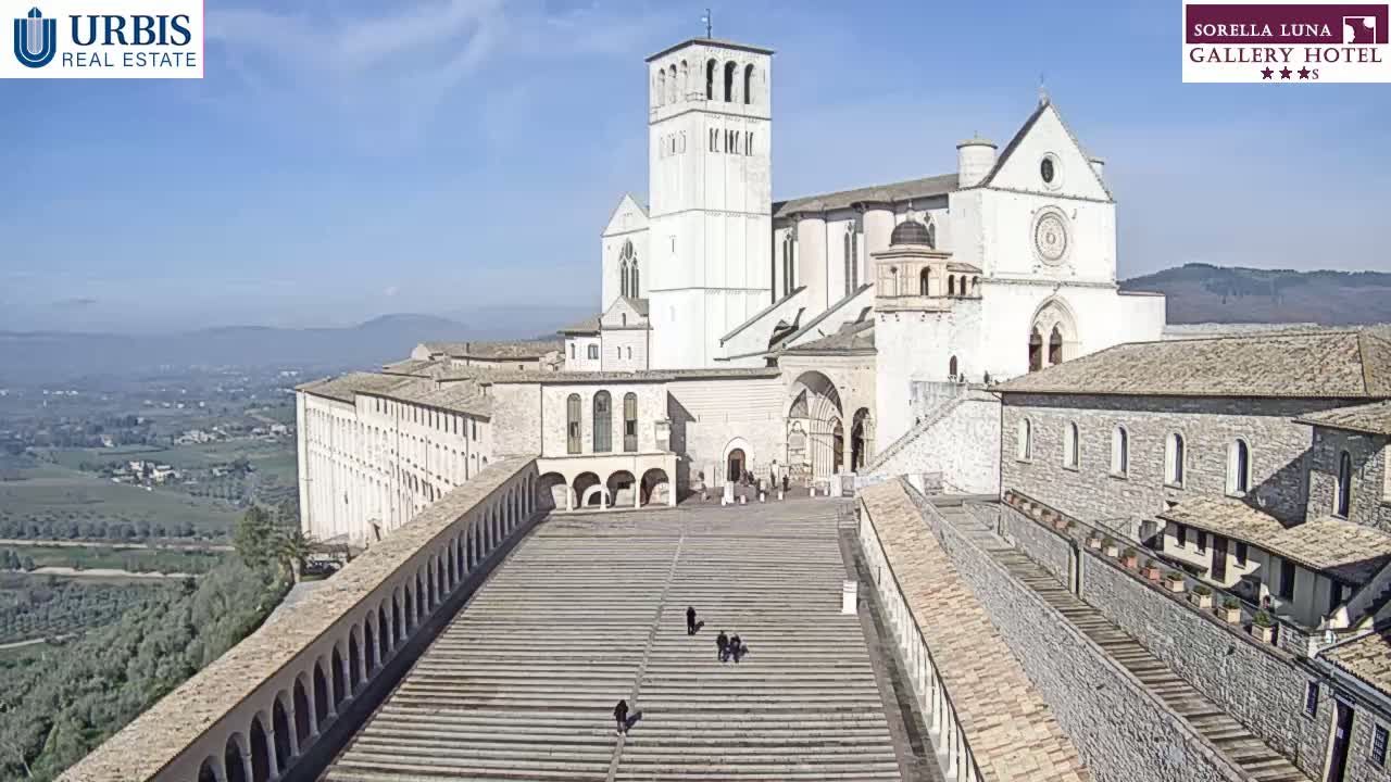 A large, light-colored basilica complex with a tall bell tower sits atop a hill overlooking a valley on a clear, sunny day.