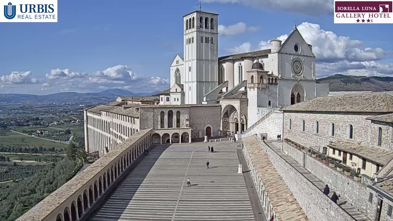 A large, light-colored basilica and adjoining buildings sit atop a hill overlooking a valley under a mostly sunny sky with a few clouds.