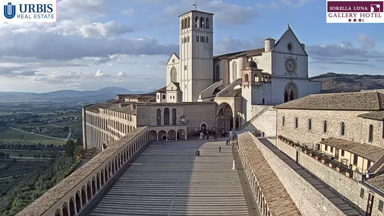 A large stone basilica and adjacent buildings are viewed from above on a partly cloudy day, overlooking a sprawling landscape.