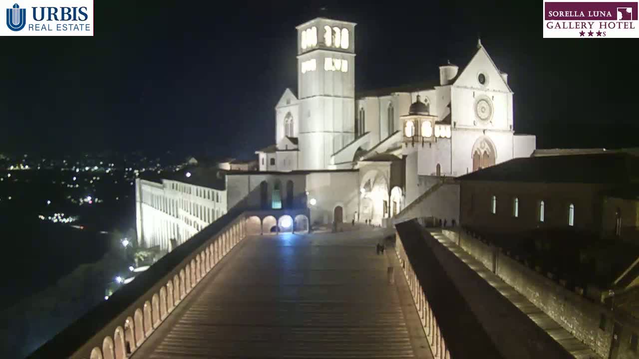 A large, illuminated basilica stands at night overlooking a city, with a long, paved walkway leading down to it from an elevated viewpoint.