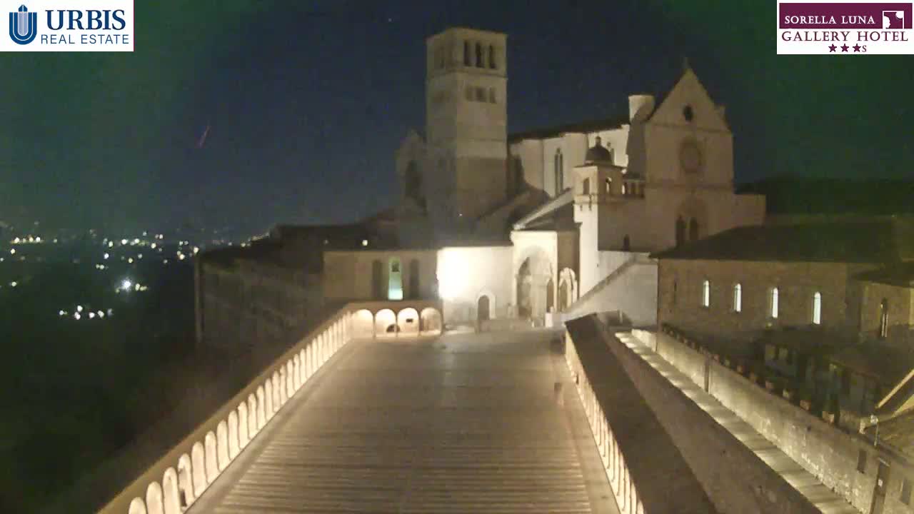 A nighttime view of a large stone building complex, illuminated, with a long, descending walkway in the foreground and a distant city glowing faintly under a clear, dark sky.