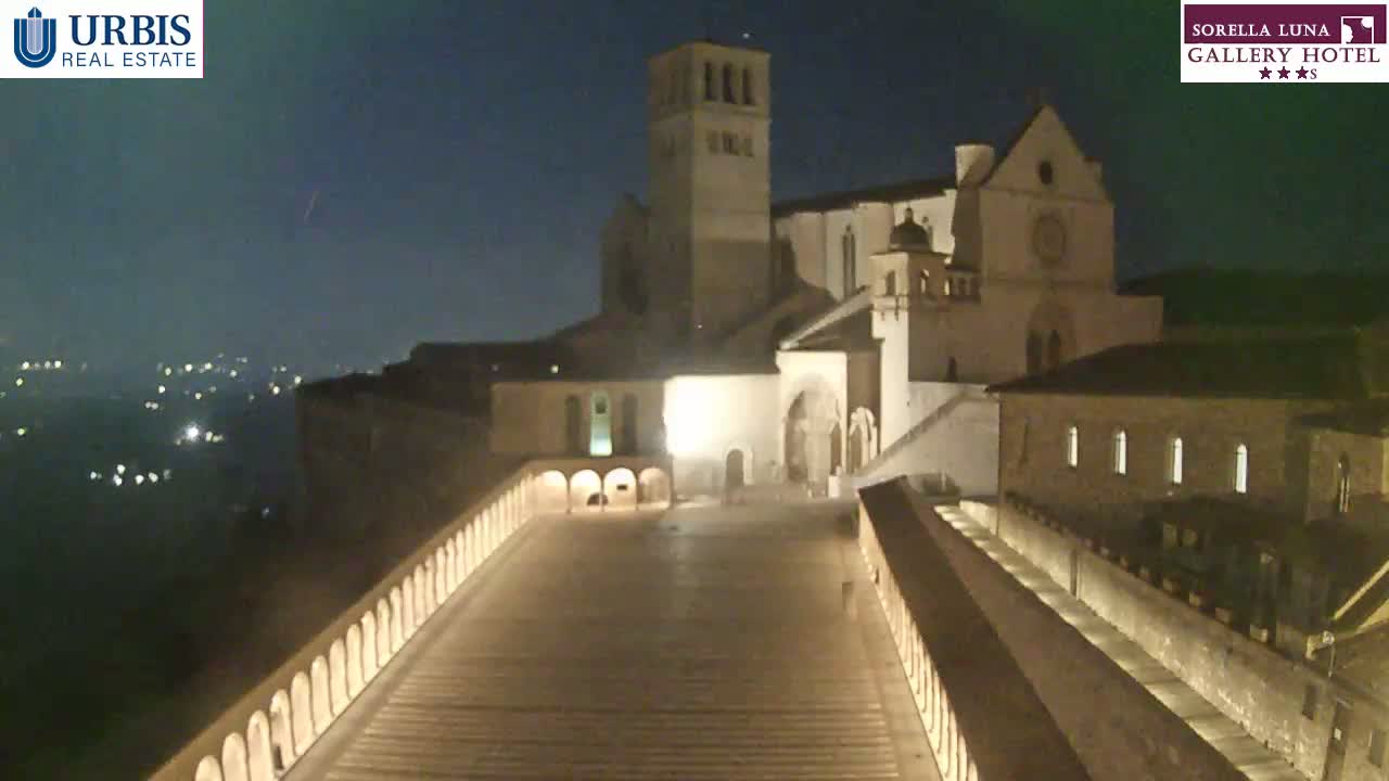 A large, illuminated stone building complex sits atop a hill at night, overlooking a distant city under a mostly clear sky.
