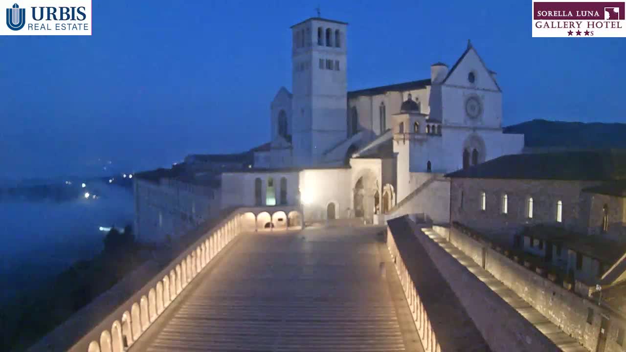 A large, light-colored stone church complex is illuminated at night, with a low-lying fog in the valley below under a twilight sky.