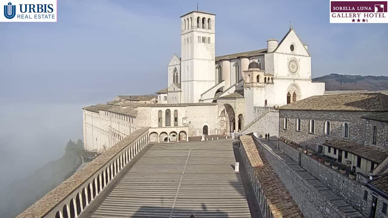 A large, light-colored basilica sits atop a hill shrouded in low-lying fog under a clear blue sky.
