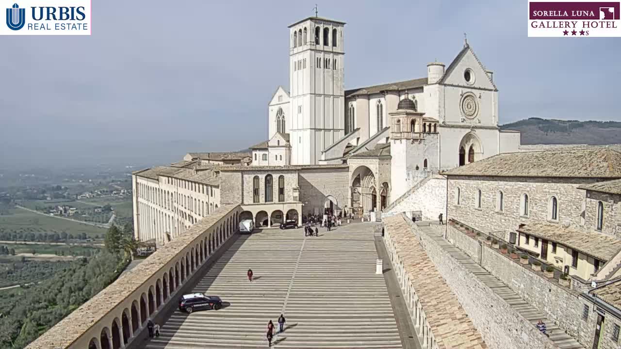 A large, light-colored basilica and adjacent buildings are situated on a hill overlooking a valley, under a bright, clear sky.