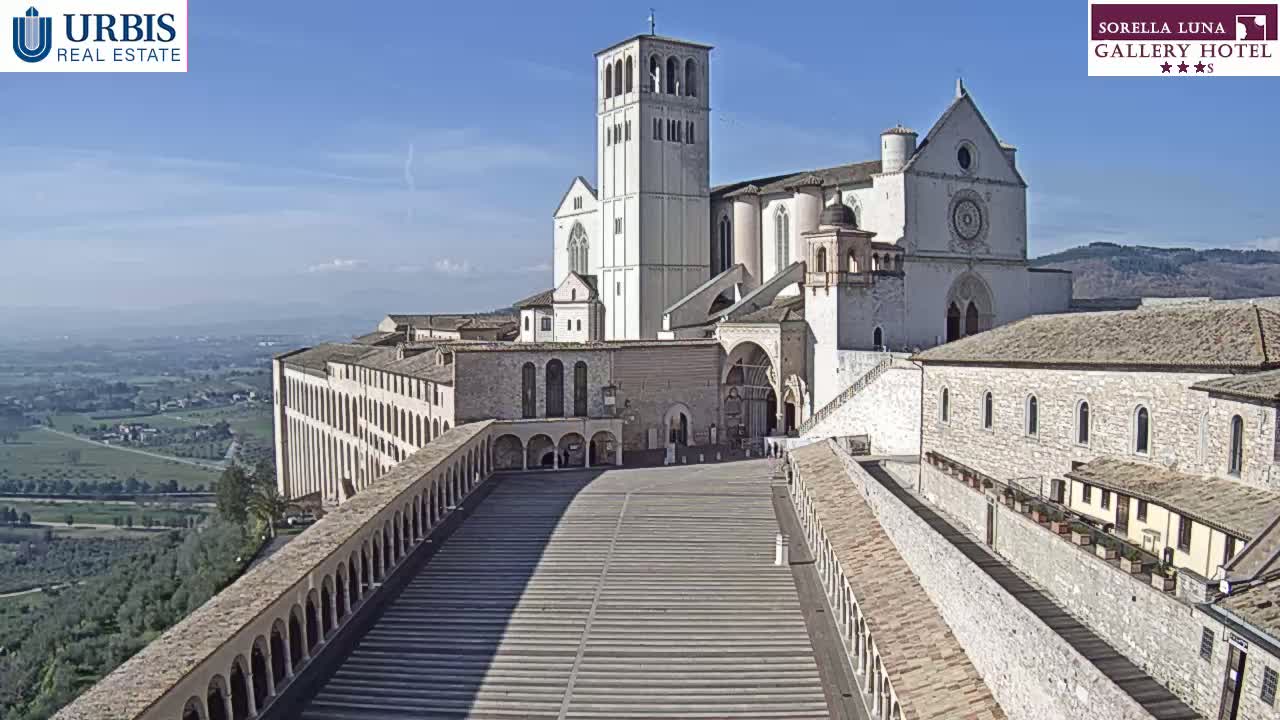 A large, light-colored stone basilica and adjoining buildings stand on a hilltop overlooking a valley under a clear blue sky.