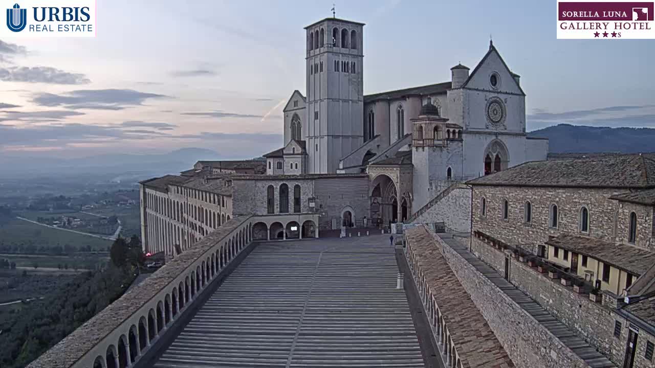 A large stone basilica sits atop a hill overlooking a valley at dawn, under a pale sky.