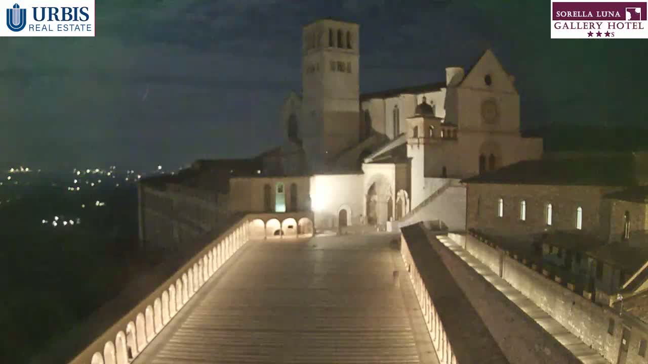 A large, illuminated stone building complex is shown at night, overlooking a valley with distant lights.