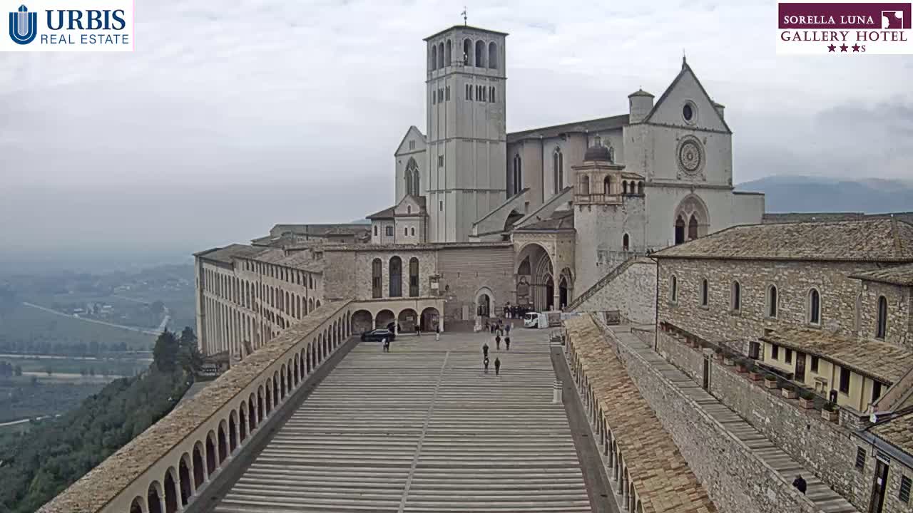 A large stone basilica and surrounding buildings are seen on an overcast day, with a long, wide staircase leading to the main structure, overlooking a valley.