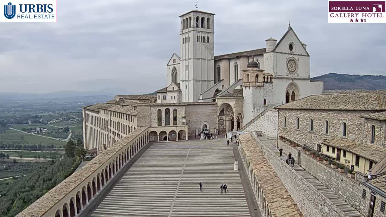 A large stone basilica and adjoining buildings are situated on a hill overlooking a valley under an overcast sky.