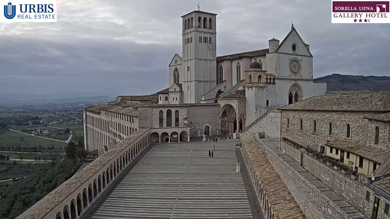 An expansive stone plaza stretches before a large basilica, with a long, low building extending to the left, all under an overcast sky.