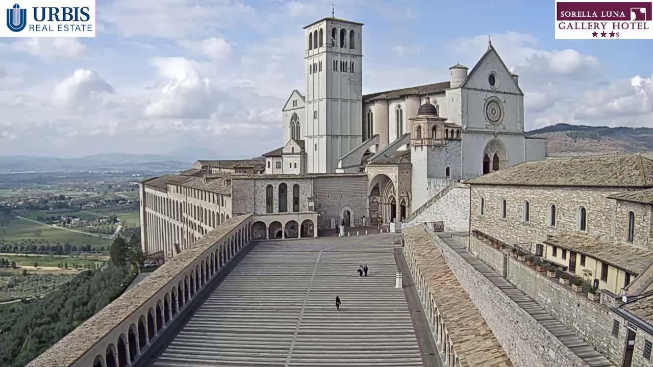 A large, light-colored stone basilica complex overlooks a sprawling landscape under a partly cloudy sky.