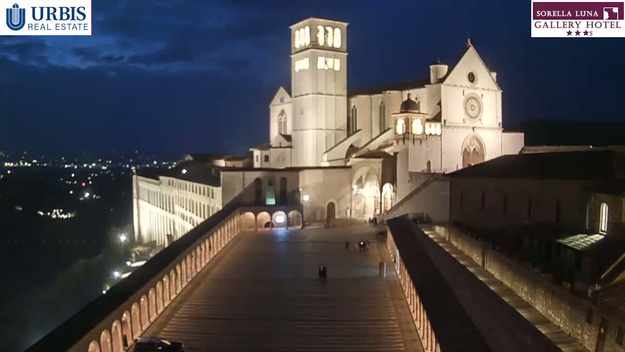 A large, illuminated basilica sits atop a hill at night, overlooking a city with a long, arcaded pathway leading to its entrance under a dark, mostly clear sky.