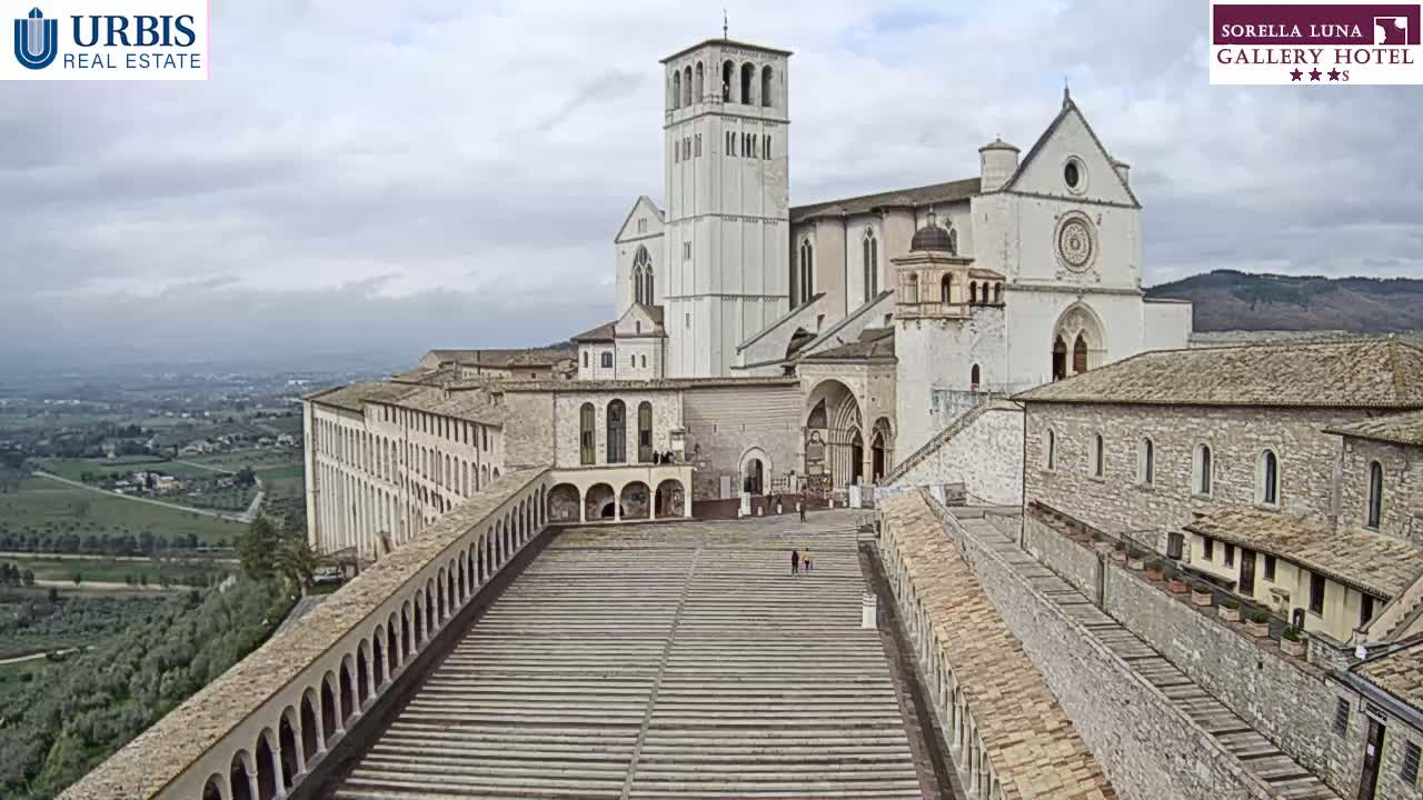 A large, light-colored stone basilica complex with attached buildings is viewed from above on an overcast day, overlooking a wide expanse of rolling hills and farmland.