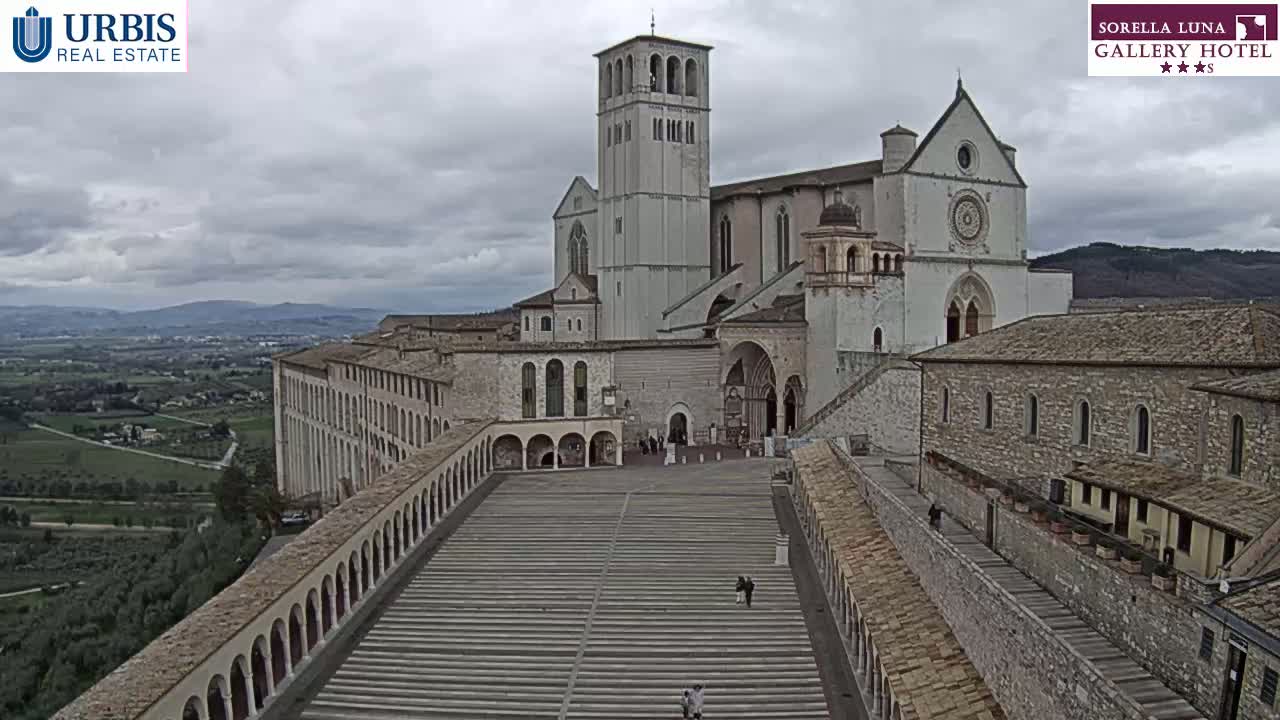 A large, light-colored basilica sits atop a hill overlooking a valley under a cloudy sky.