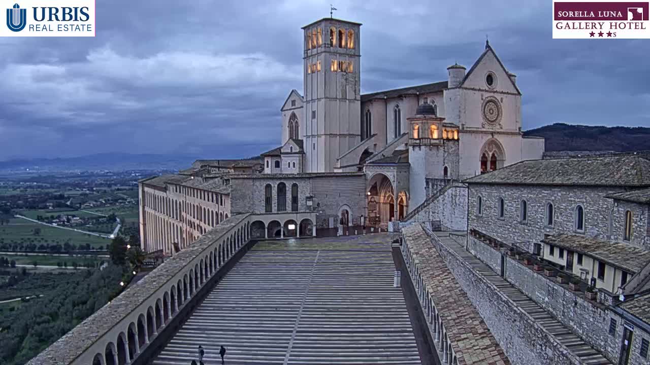 A large stone basilica and adjoining buildings are situated on a hill overlooking a valley under a cloudy twilight sky.
