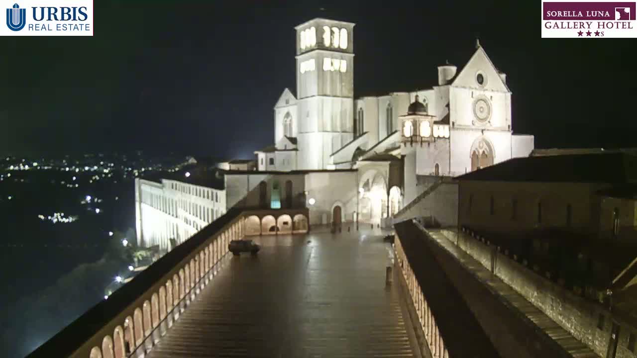 A large, illuminated basilica stands at night, overlooking a city with a paved plaza and colonnaded walkways, under a clear, dark sky.