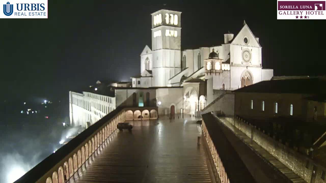 A large, illuminated basilica is seen at night in the rain, viewed from a high vantage point across a wet, paved walkway flanked by balustrades.