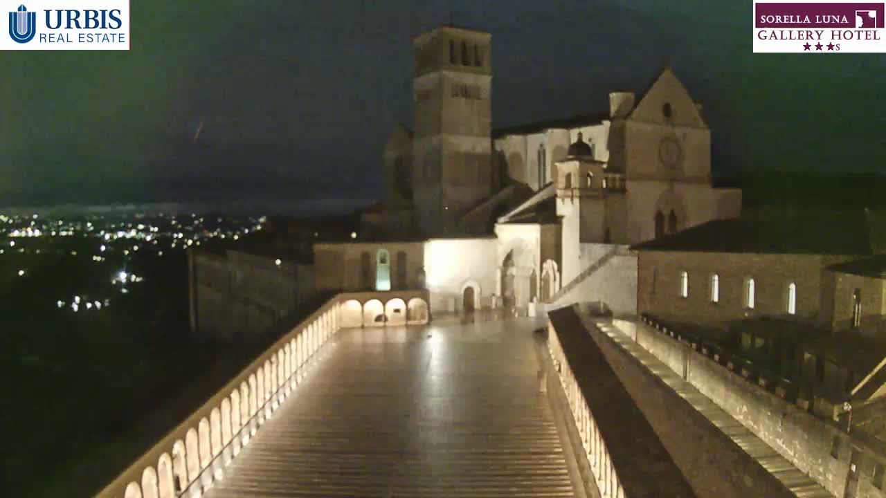 A large, illuminated stone complex is viewed at night from above, with a city and lights visible in the distance under a dark, possibly stormy sky.