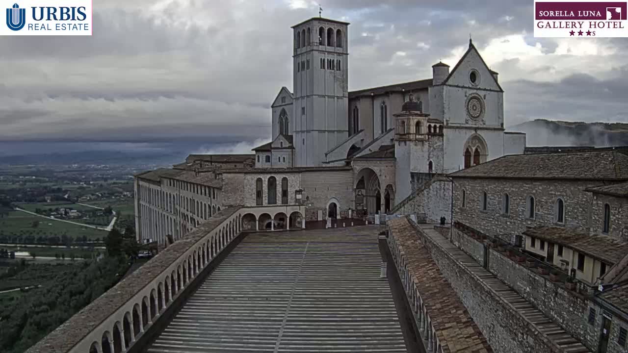 A large stone basilica and its surrounding complex sits atop a hill overlooking a valley shrouded in morning mist.