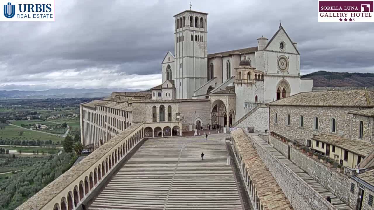 A large stone basilica and adjacent buildings are seen from an aerial perspective on an overcast day.