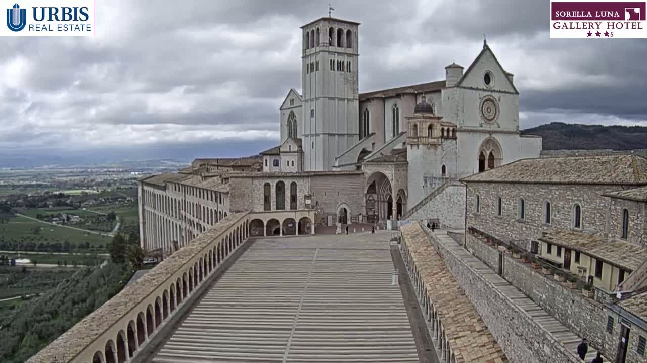 A large stone basilica complex is situated on a hill overlooking a valley under a cloudy sky.