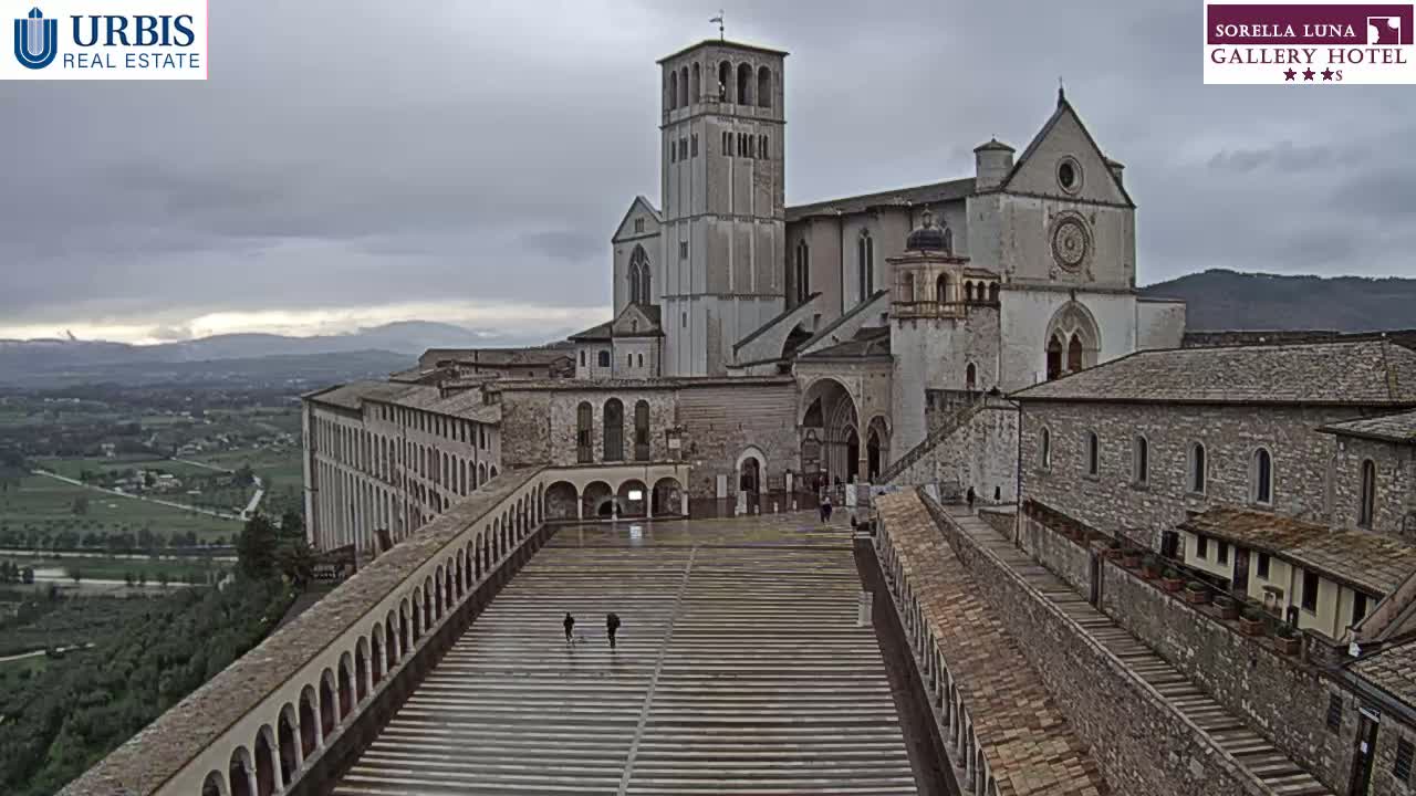 A large stone basilica and adjoining buildings overlook a sprawling valley under a cloudy sky.