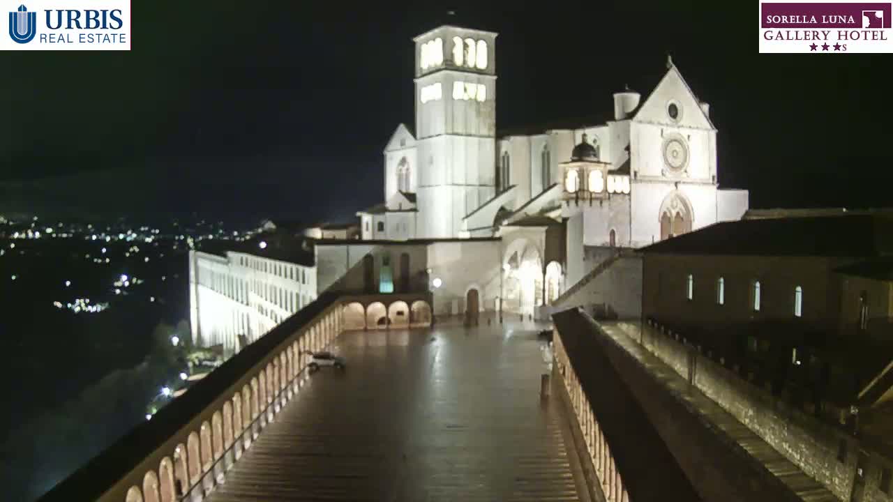 A large, illuminated basilica stands at night, overlooked by a long, arched walkway, with a city visible in the distance under a clear, dark sky.