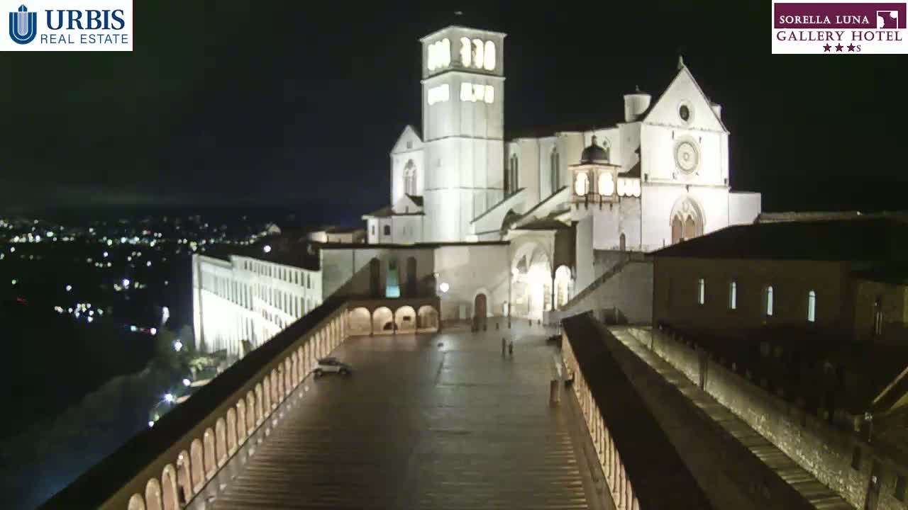 A large, illuminated basilica stands at night overlooking a city with a plaza in front.