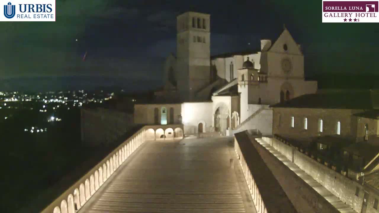 A large, illuminated stone structure, possibly a church, is visible at night, with a city's lights in the distance under a mostly clear sky.