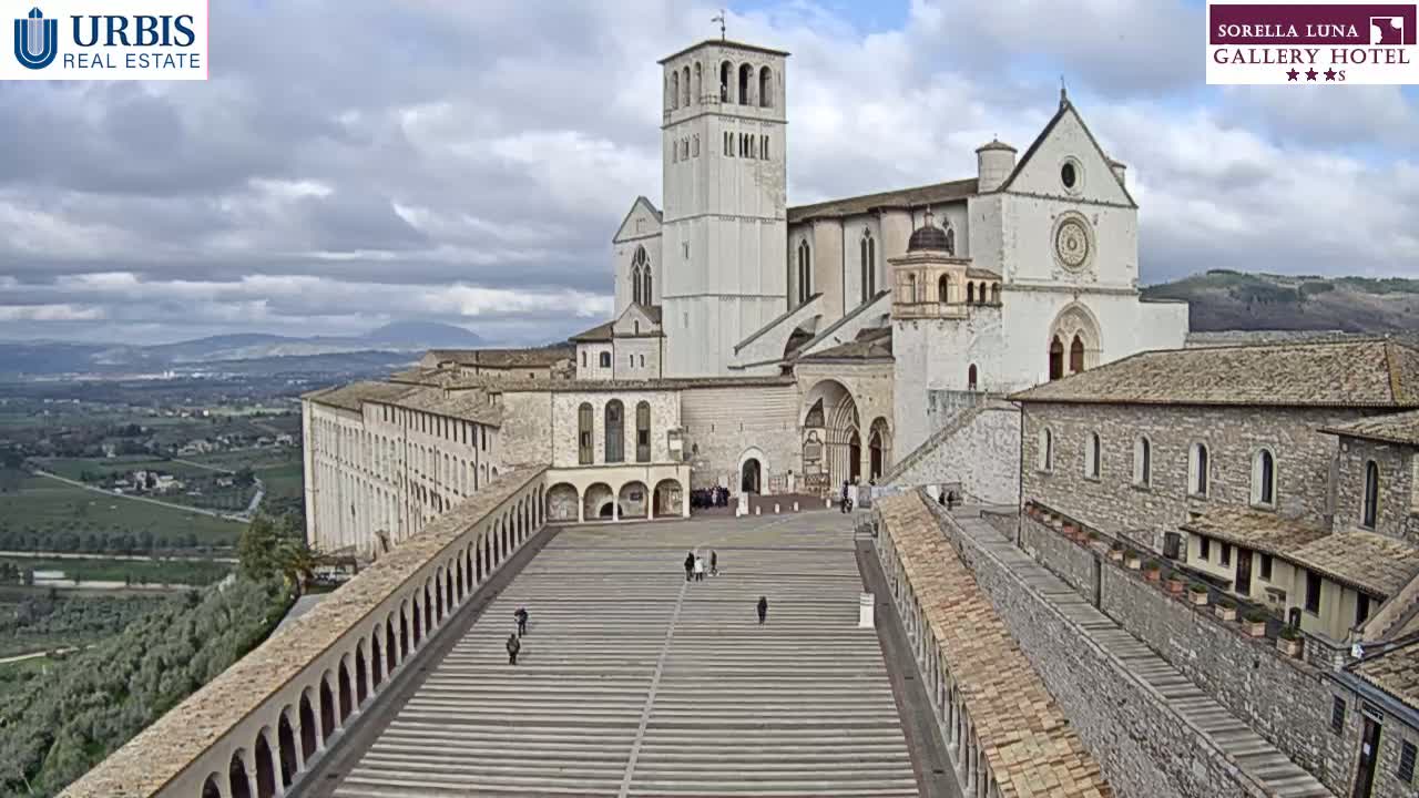 A large stone basilica and surrounding buildings are viewed from above on an overcast day, with a paved plaza separating the structures and a valley visible in the distance.