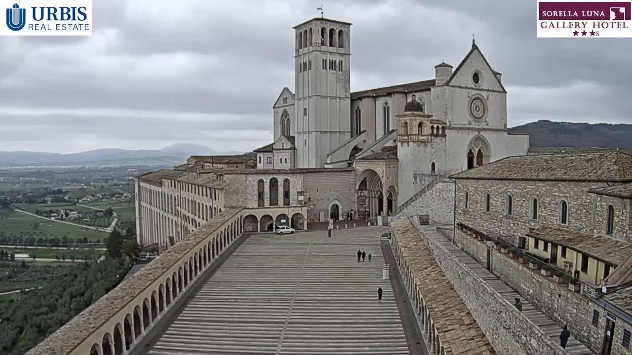 A large stone basilica and adjoining buildings are viewed from above on an overcast day, with a spacious plaza in the foreground.