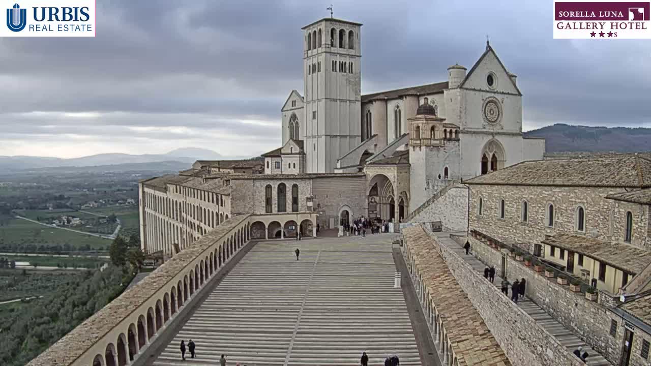 A large stone basilica and adjoining buildings stand on a hill overlooking a sprawling landscape under a cloudy sky.
