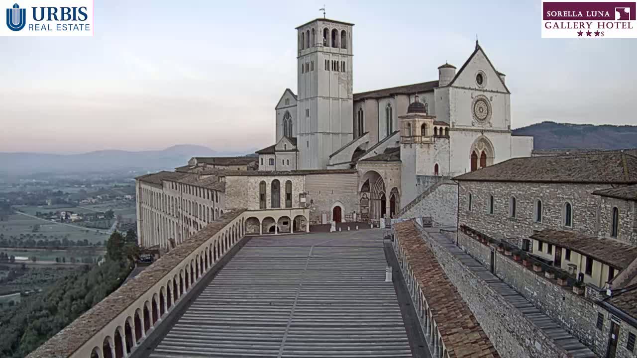 A large stone basilica and adjacent buildings overlook a wide, paved plaza under a pale, clear sky.