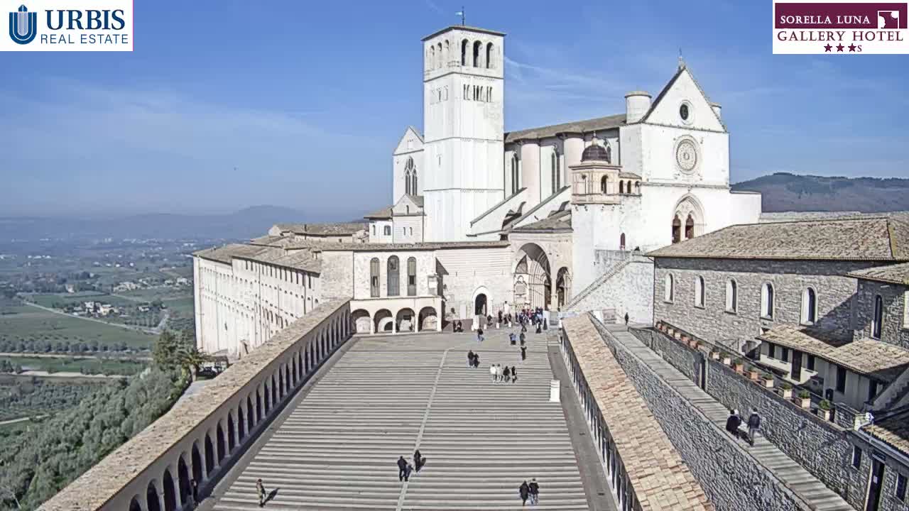 A large, light-colored basilica and adjoining buildings are situated on a hill overlooking a valley on a sunny day.