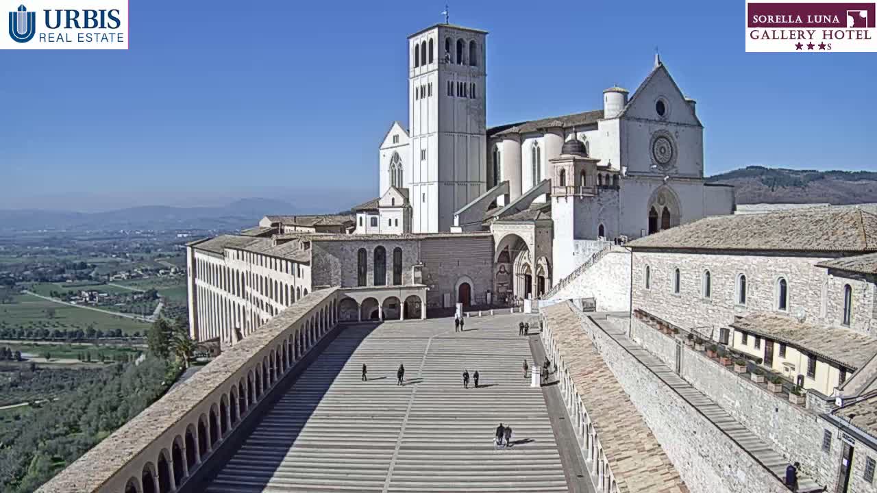 A large, light-colored stone basilica complex sits on a hilltop under a clear blue sky, overlooking a sprawling landscape.